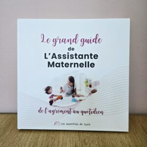 Photo de la couverture du livre rigide premium Le Grand Guide de l’Assistante Maternelle, posé debout sur une table en bois avec fond rose.