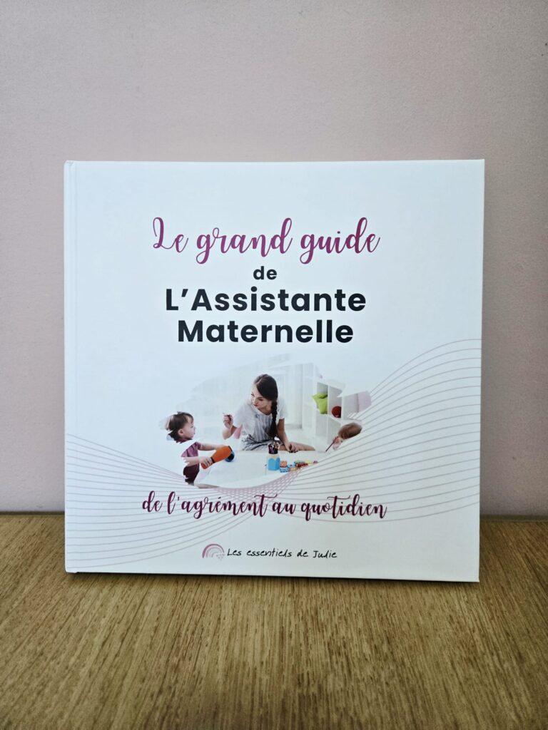 Photo de la couverture du livre rigide premium Le Grand Guide de l’Assistante Maternelle, posé debout sur une table en bois avec fond rose.