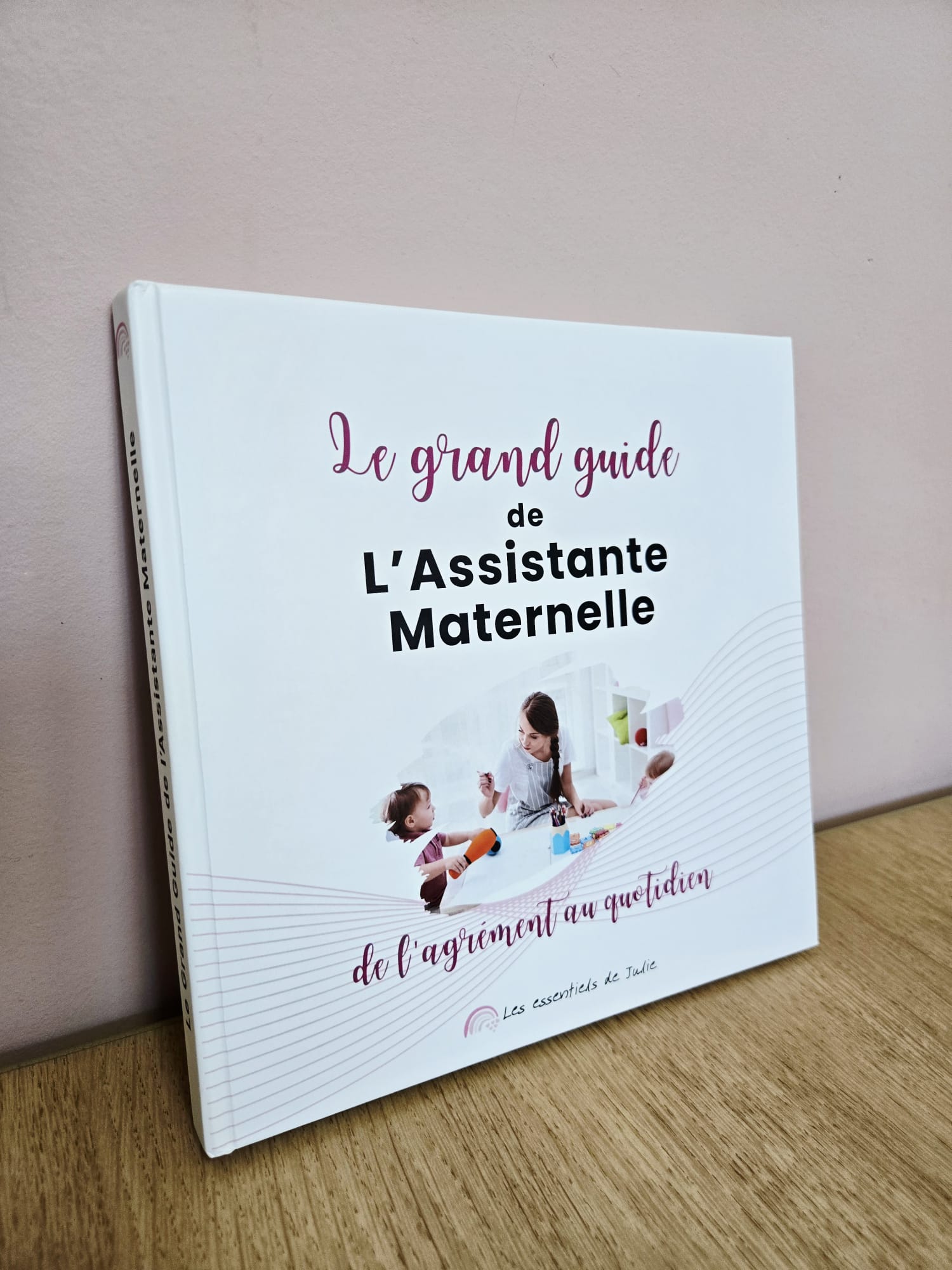 Le Grand Guide de l’Assistante Maternelle – Édition Rigide Premium 2 Photo de la version rigide premium du livre Le Grand Guide de l’Assistante Maternelle, montrant la couverture et la tranche, posée sur une table en bois devant un mur rose.